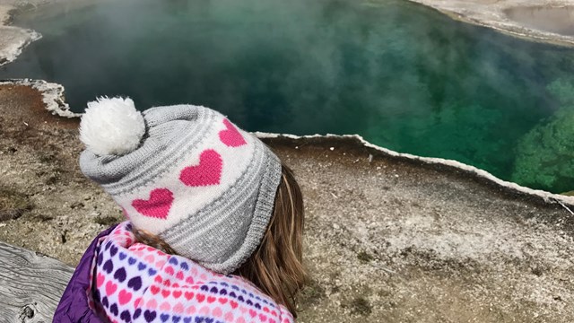 Child wearing a winter hat and coat looking out across a deep, aqua-green hot spring.