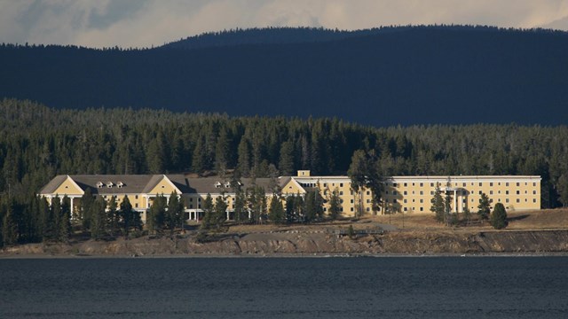 Yellow and white hotel sits in the conifer trees near the shore of a large lake.
