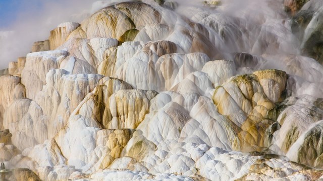 A terraced hot spring of whites, yellows, and oranges with steam rising from it.