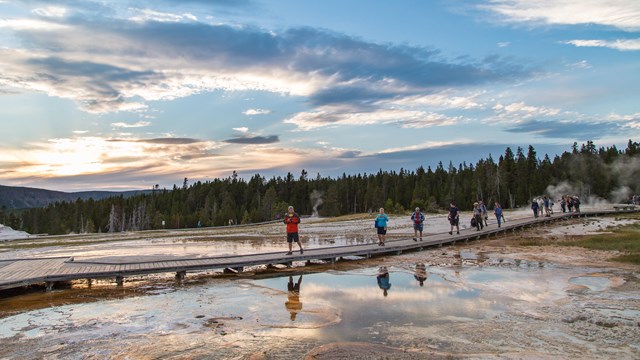 People walk across a boardwalk leading across a hillside covered in steaming hot springs.