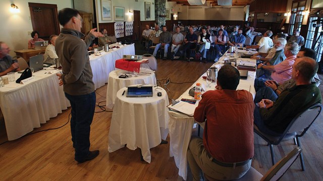 A presenter speaks in front of an audience of multiple agency representatives.