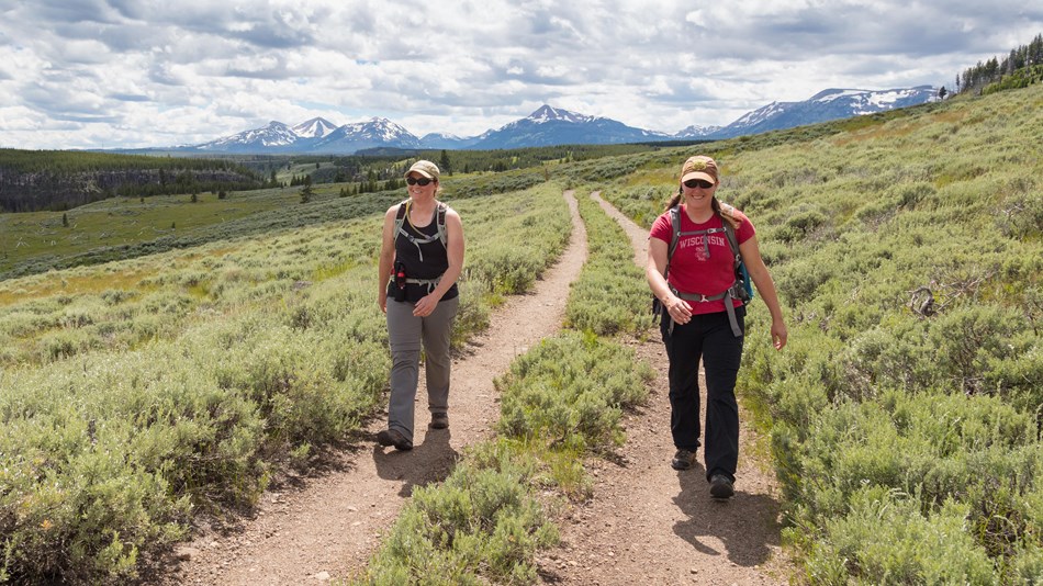 Two women walking on a trail with mountains in the landscape in the background.