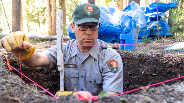 A uniformed man stands in a pit using twine to measure the pit.