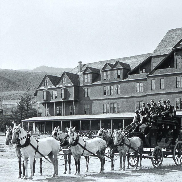 Black and white photo of a six horse tallyho stagecoach full of people is stopped in front of hotel