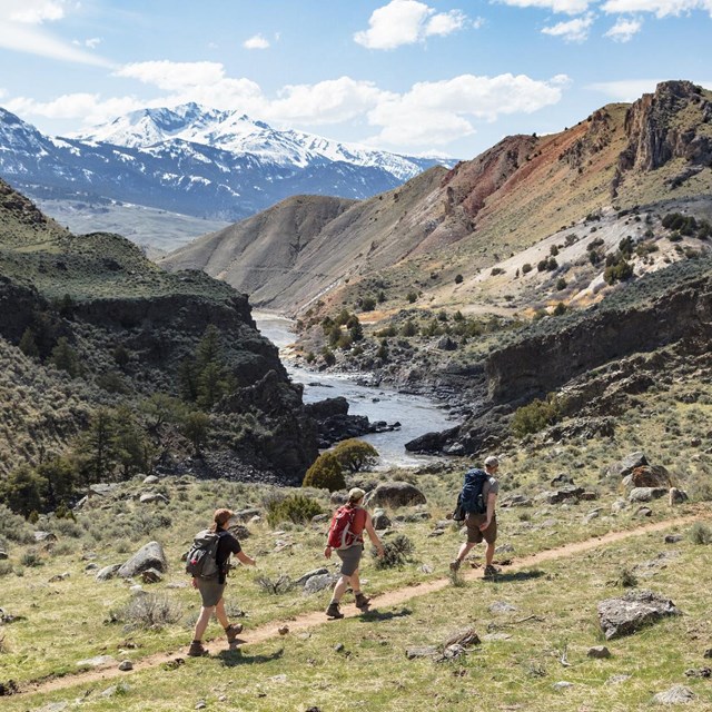 Hikers walking along a trail with river and mountain views.