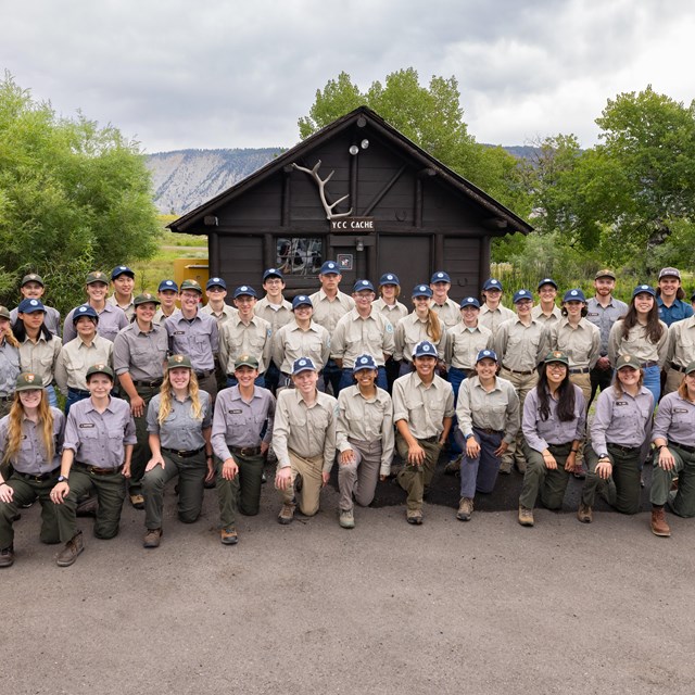 a group photo of people standing in front of a small log cabin building