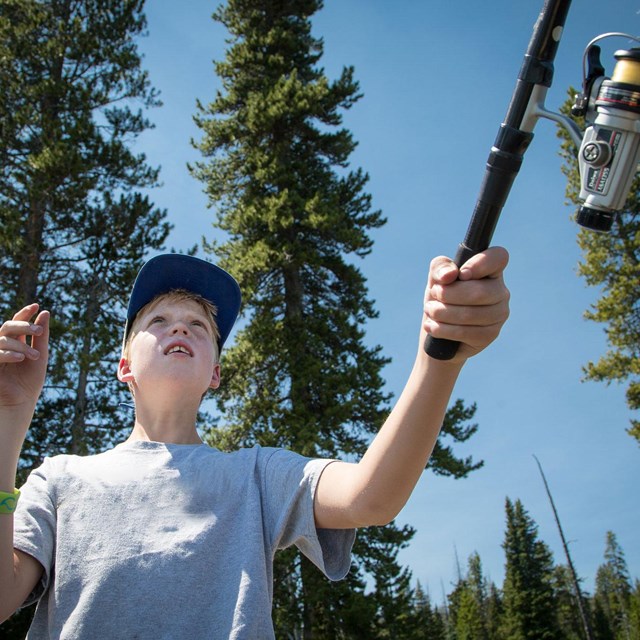 A boy with a hat on casts a fishing rod and reel in front of tall conifer trees.