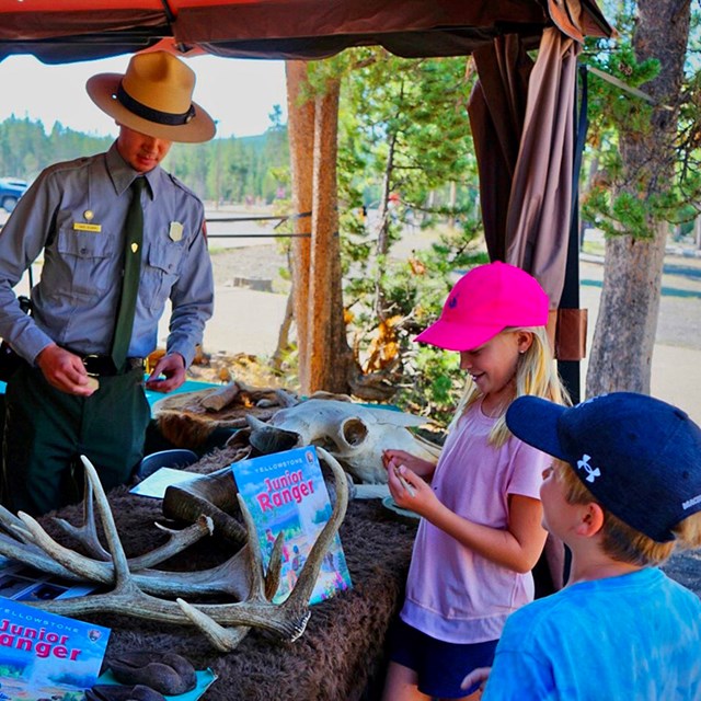 Two school-aged children viewing elk antlers and Junior Ranger booklets and speaking to two park ran