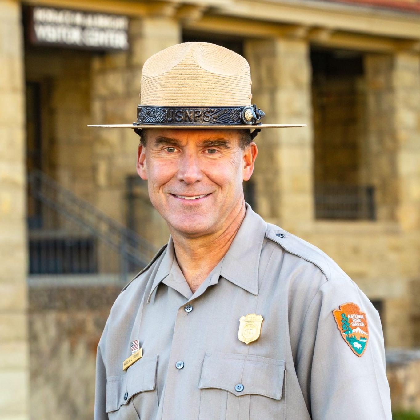 A smiling male park ranger in uniform standing in front a historic stone building