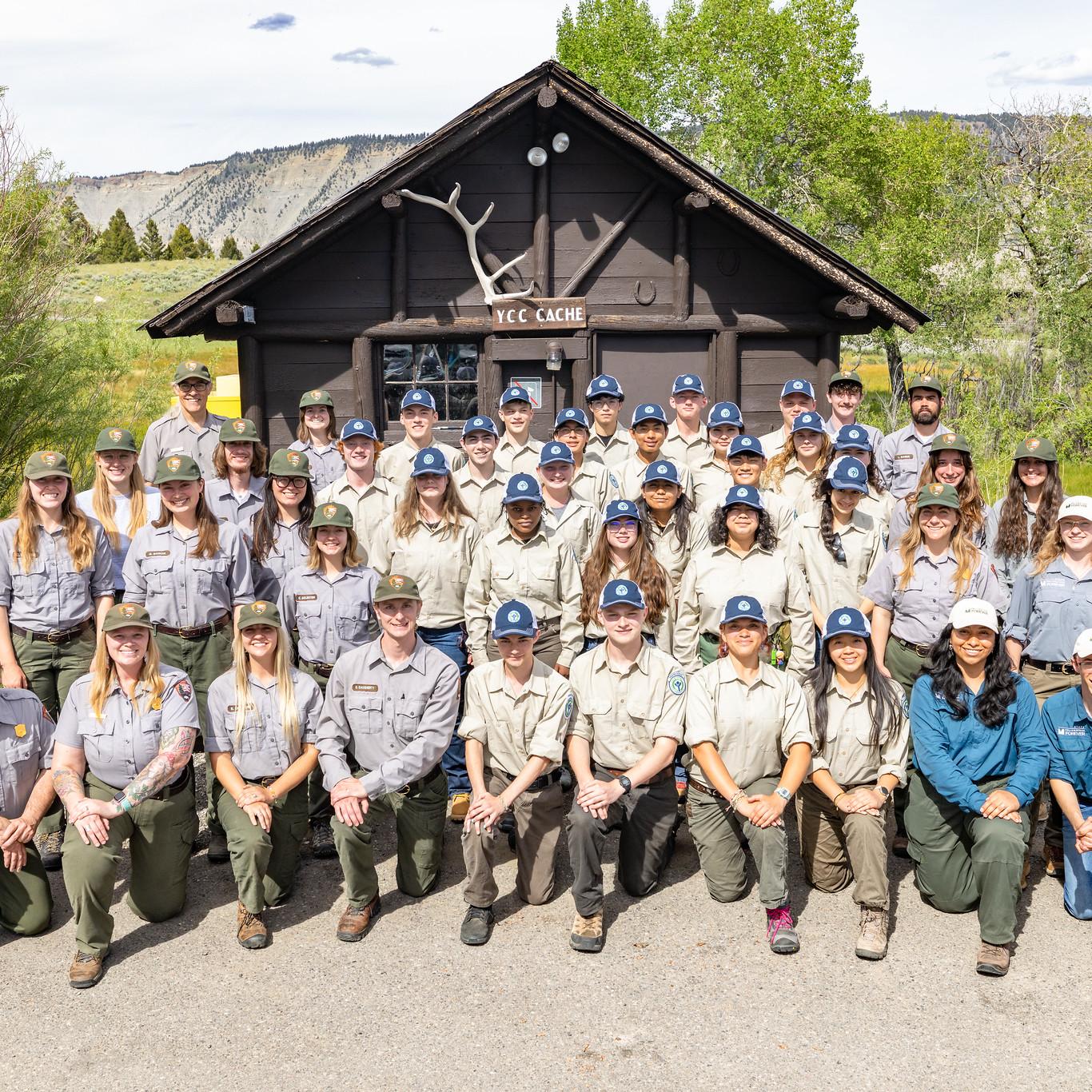 A group of teens and young adults posing and smiling in front of a small log-structure shed.
