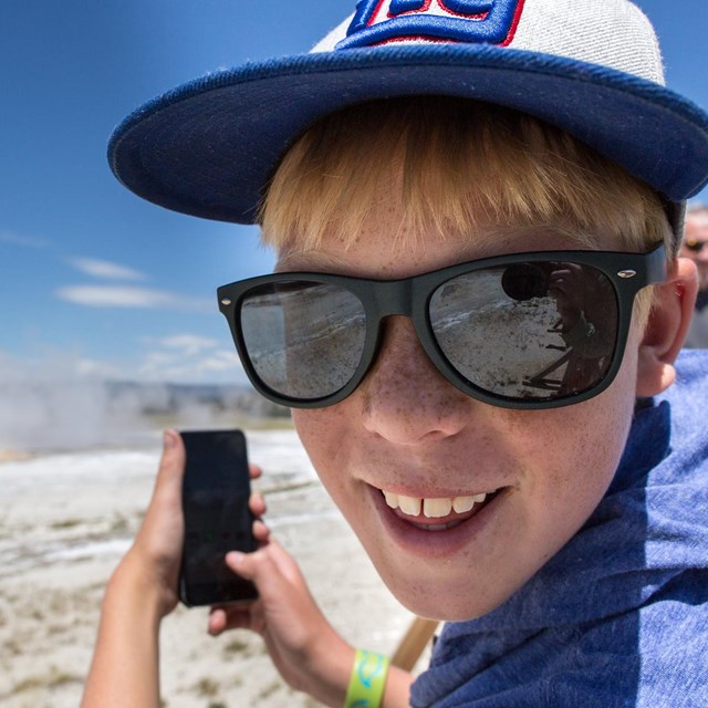 Close up of a young boy with a hat and sunglasses taking a picture of a geyser with a phone.