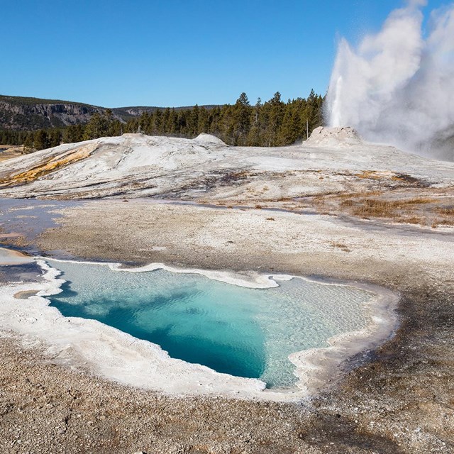 A blue hot spring with a geyser erupting in the background.