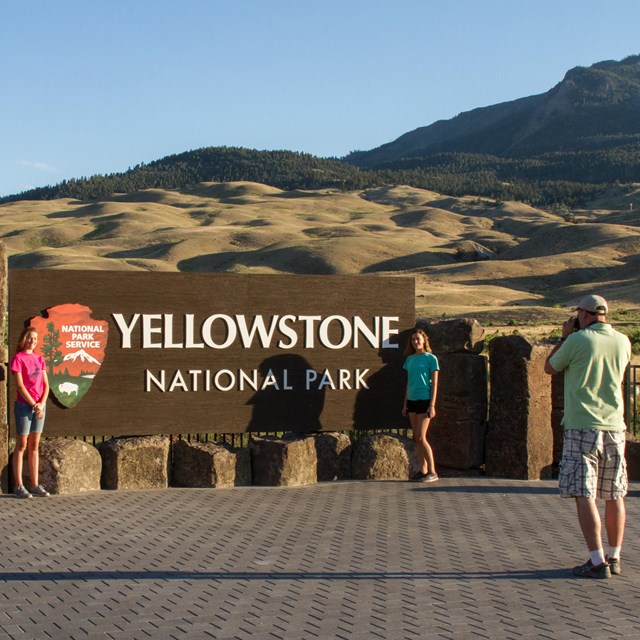 Two girls stand in front of a large Yellowstone National Park sign while their dad photographs them