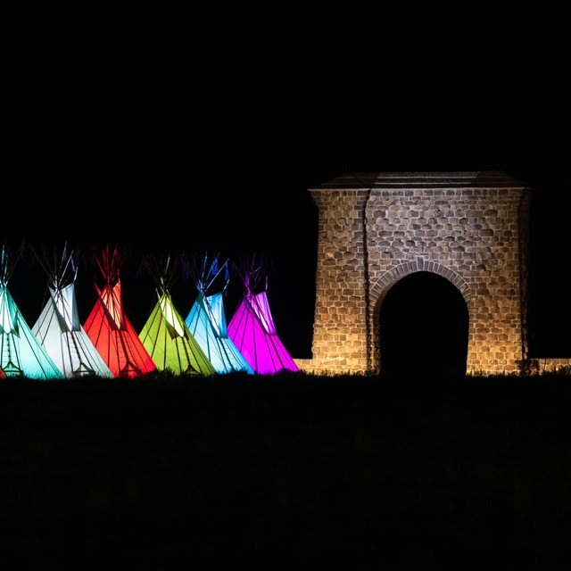 a row of colorful, illuminated teepees near a large, stone archway at night
