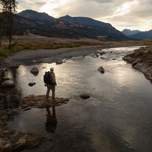 A hiker stands on a rock on the edge of a river in the fading daylight.