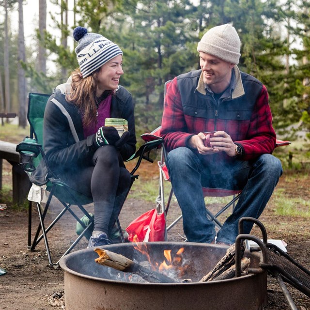 A man and a woman sit by a campfire in a campsite and drink coffee.