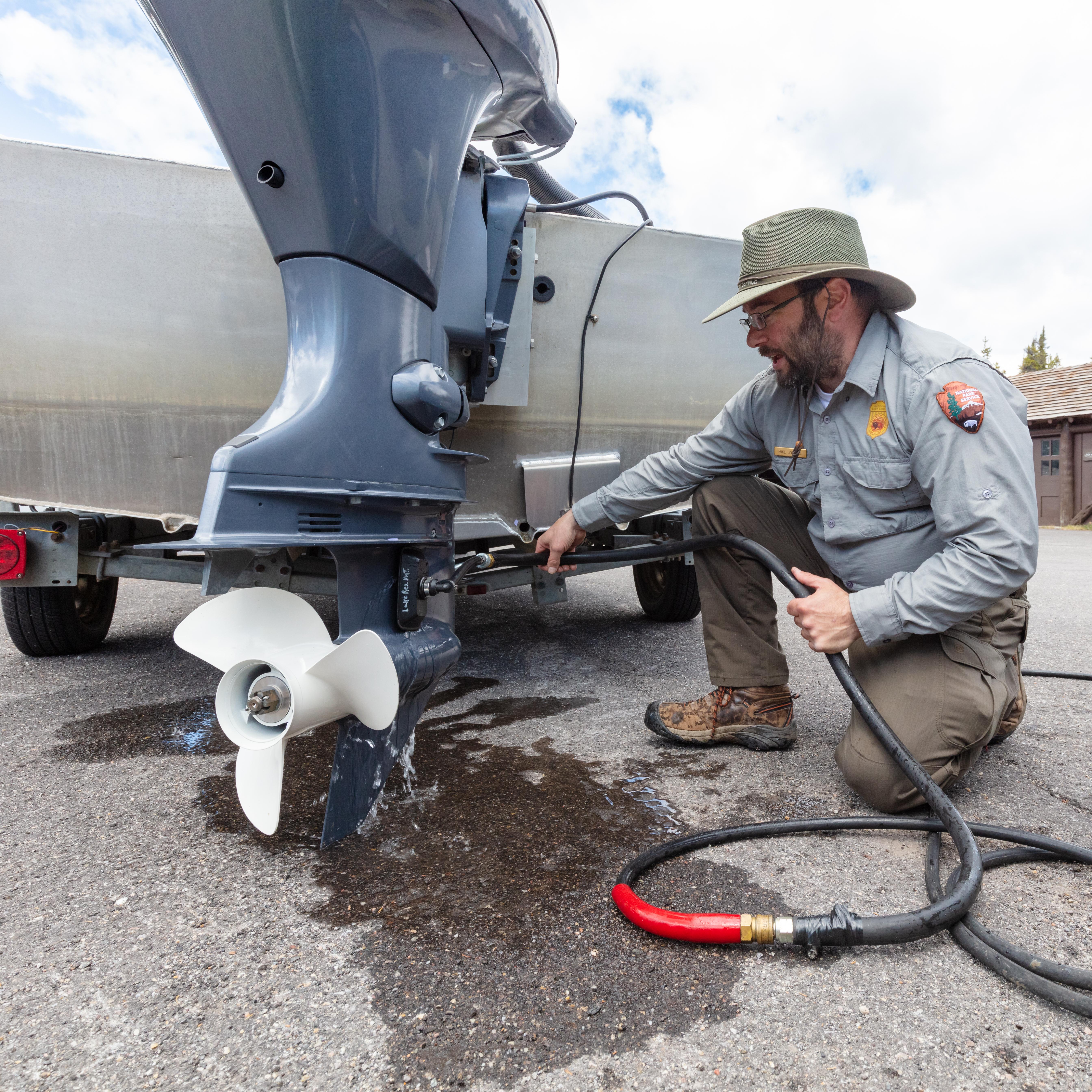 A park ranger with a dark beard and wearing a green and grey uniform kneels with a hose next a boat.