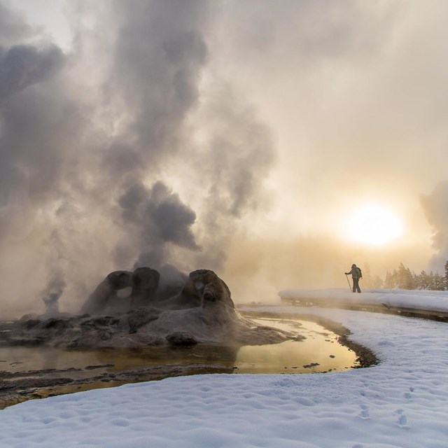 A skier enjoys a winter sunrise at near a steaming geyser.