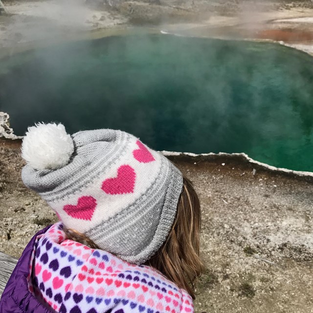 Child wearing a winter hat and coat looking out across a deep, aqua-green hot spring.
