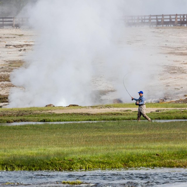 a person fly fishing from the bank of a river near a thermal area