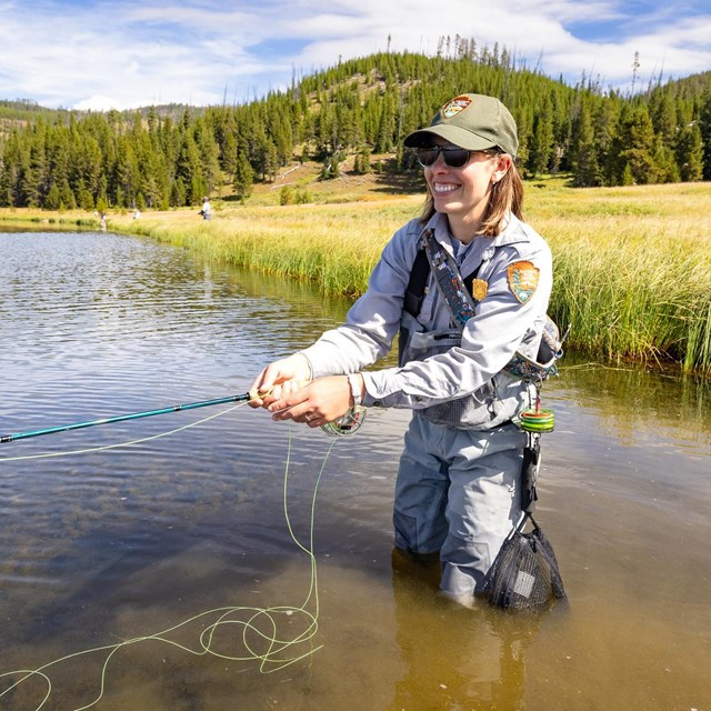 a woman wearing a park ranger uniform and waders standing in a lake with a fly fishing rod
