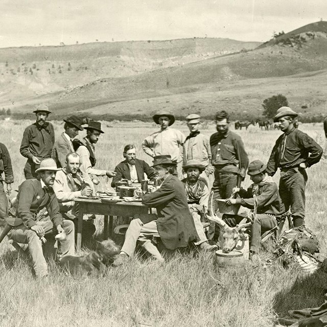 9 MEN STANDING, 6 MEN SEATED AROUND TABLE, COOKSTOVE ON GROUND, COVERED WAGON, HORSES IN BACKGROUND