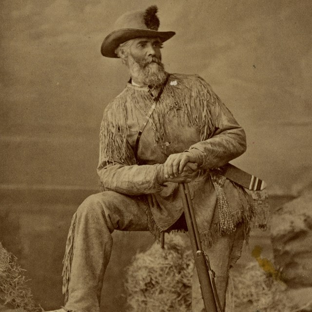 SEPIA VIEW OF MAN IN STUDIO POSE, DRESSED IN FRINGED BUCKSKINS, HAT WITH FEATHER 