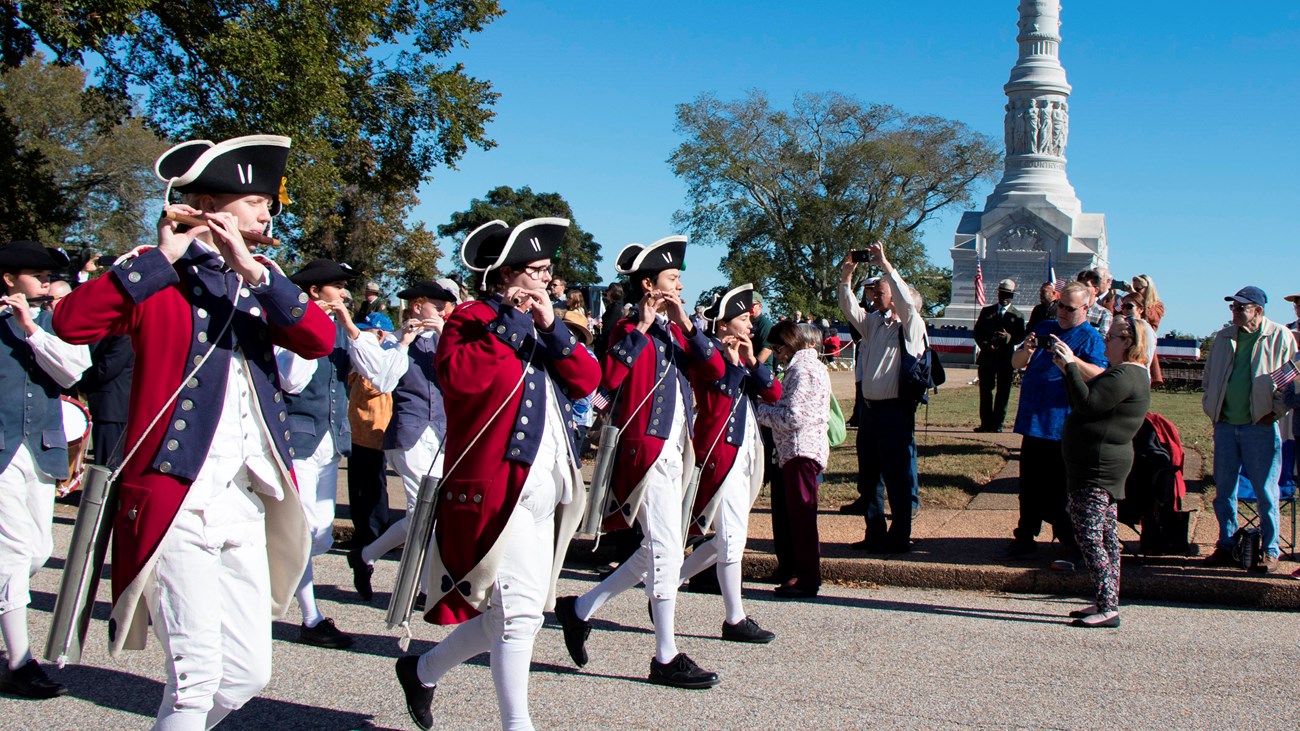 Fife and Drum wearing Red jackets play flutes in front of Victory Monument