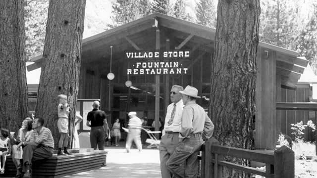 Park visitors stand and walk around a plaza in front of a store entrance; mature trees decorate the 
