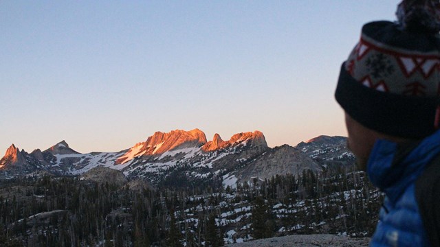 A hiker in warm jacket and hat overlooks high country scenery at sunset