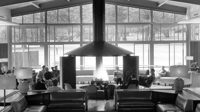 A lounge with a fireplace at center and ample lounge furniture; the high ceiling has exposed beams a