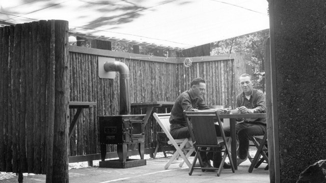 Two men sit at a round table underneath a canvas shade; the paved area is partially fenced in