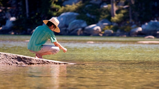 A person in a wide hat crouches on the banks of a river.