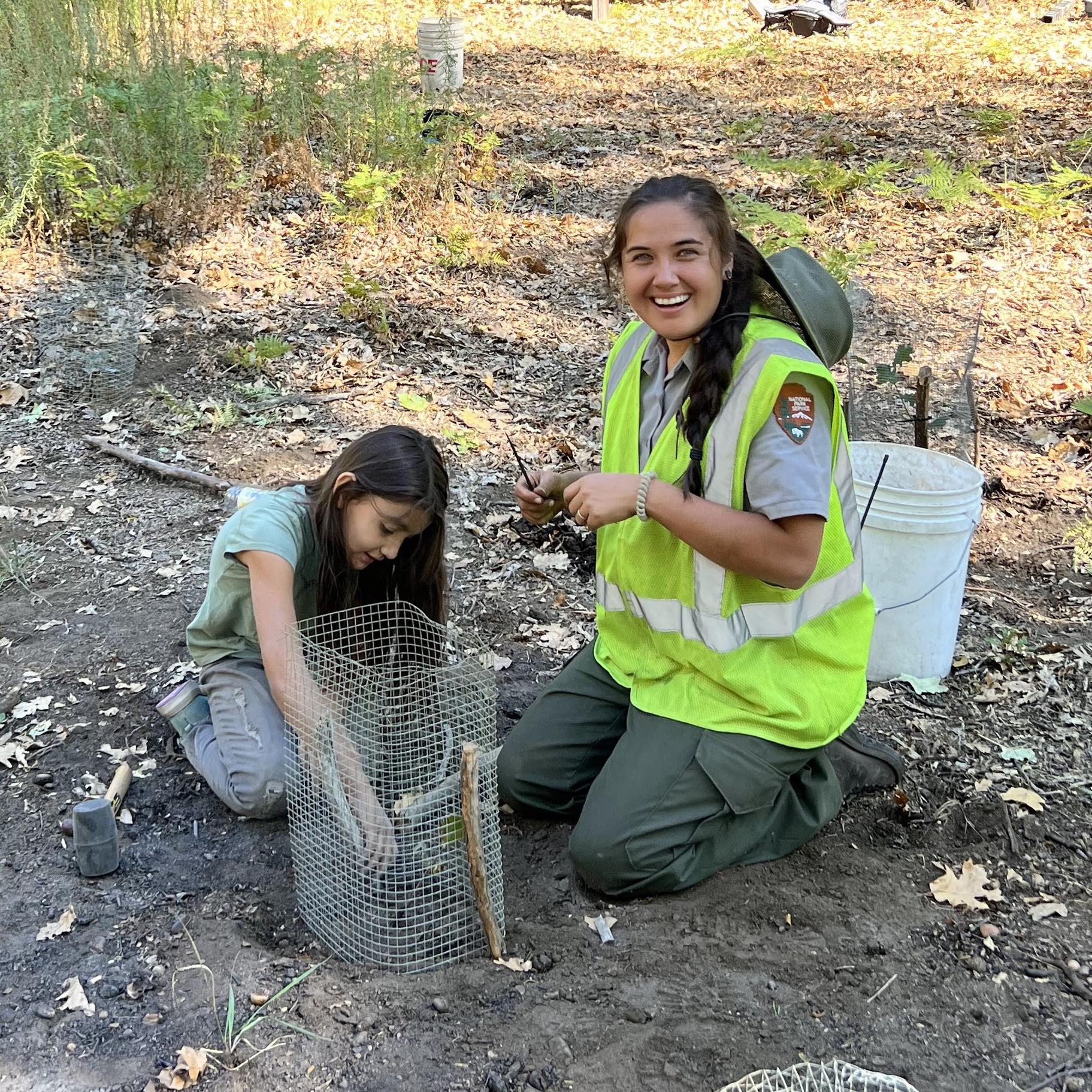 A young person and a woman kneel by a sapling in a sapling cage.