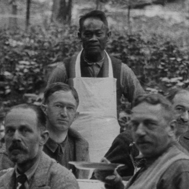Chef Tie Sing in an apron surrounded by a mountain party of men in the wilderness