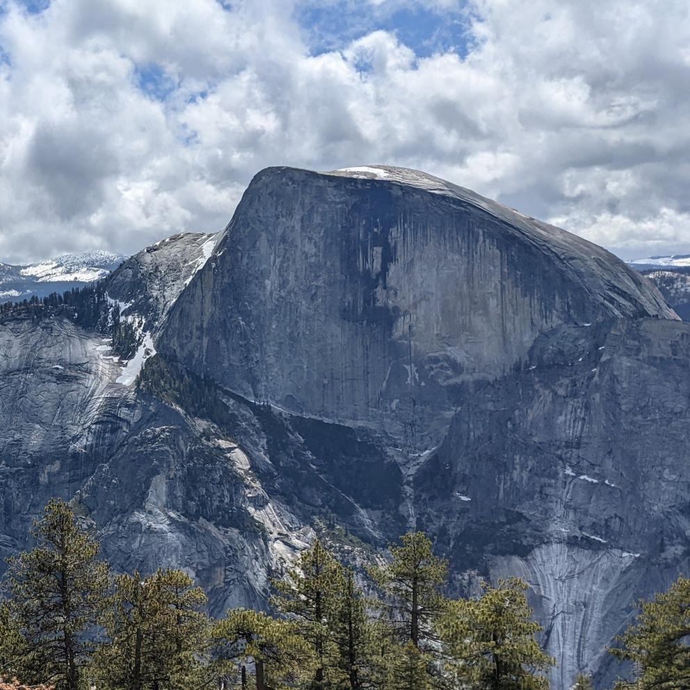 A wide picture of a sheer mountainside with clouds and trees. 