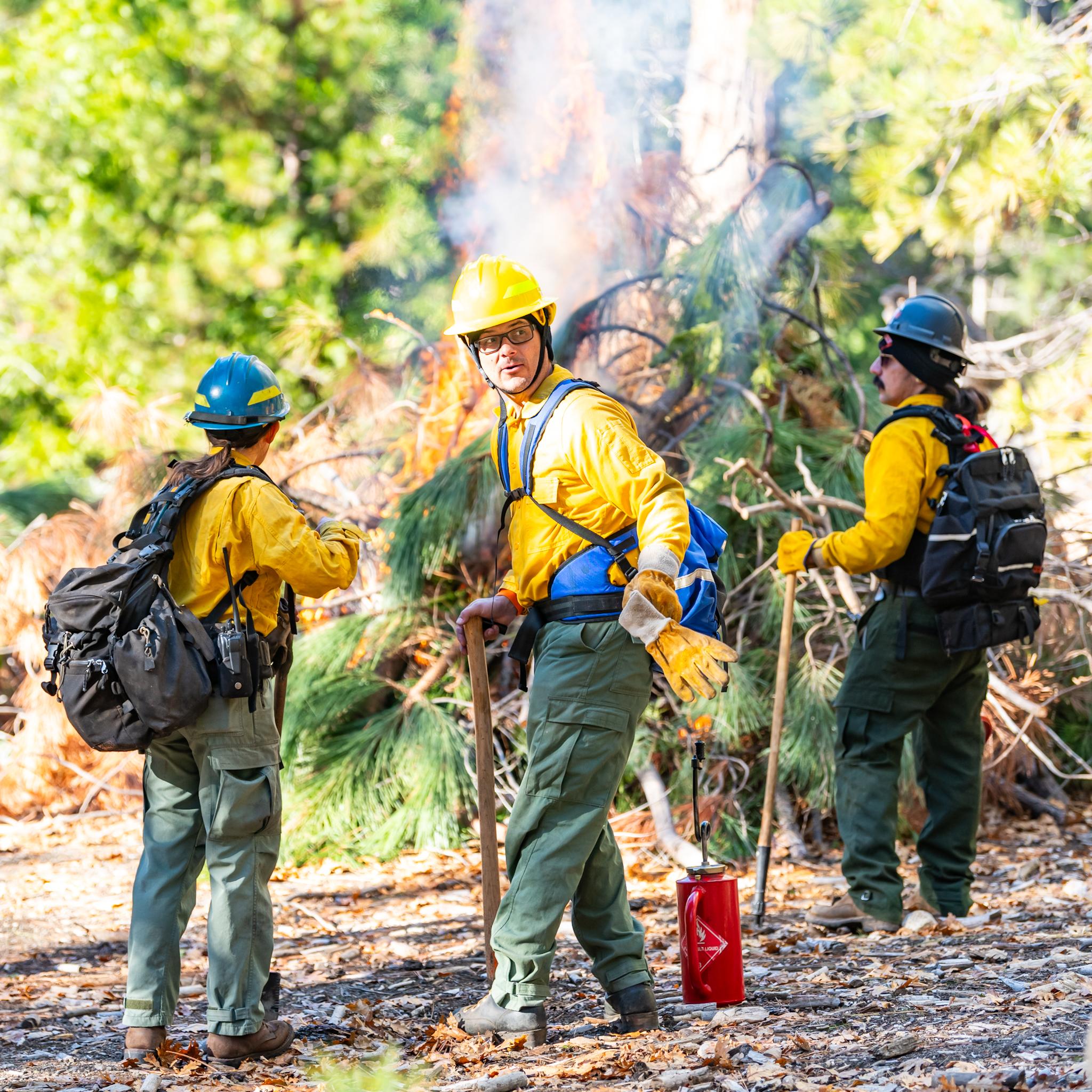 Three people wearing yellow jackets and hard hats monitor a pile burn.
