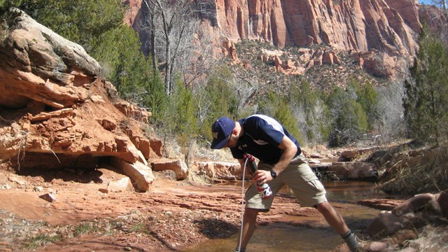 A man in a hat filtering water from a creek in front of mountains.