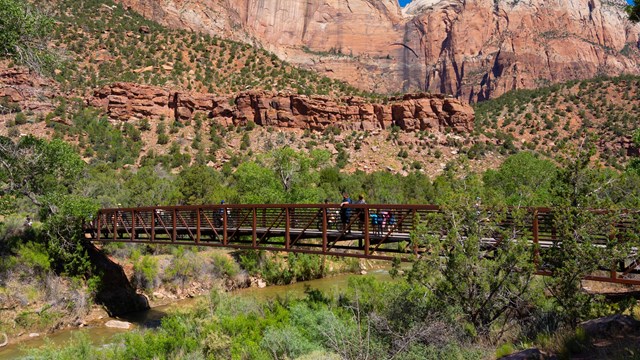 A group of people walk or bike across a bridge. In the background are huge sandstone cliffs.