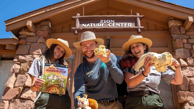 Three rangers stand in front of the Nature Center and show off various props.