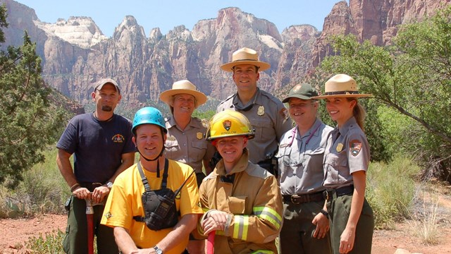 Rangers in different uniforms stand in Zion National Park surrounded by high rock walls and trees.