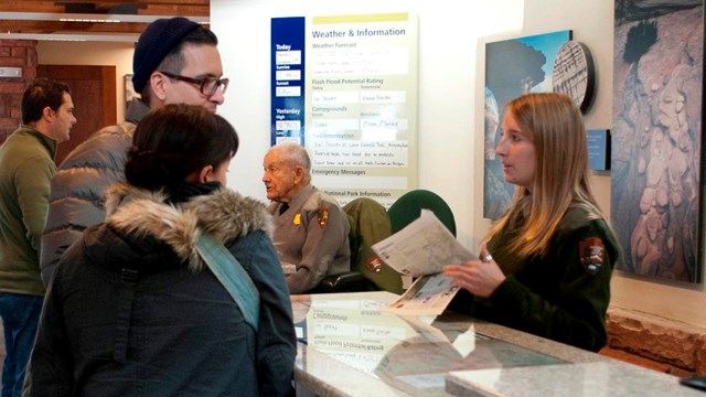 Ranger stands behind visitor center desk and speaks with visitors.