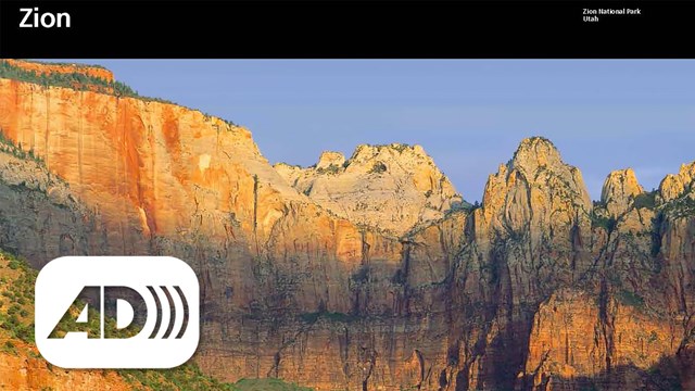 Zion unigrid with blue sky over sandstone mountains. Audio description symbol in corner. 