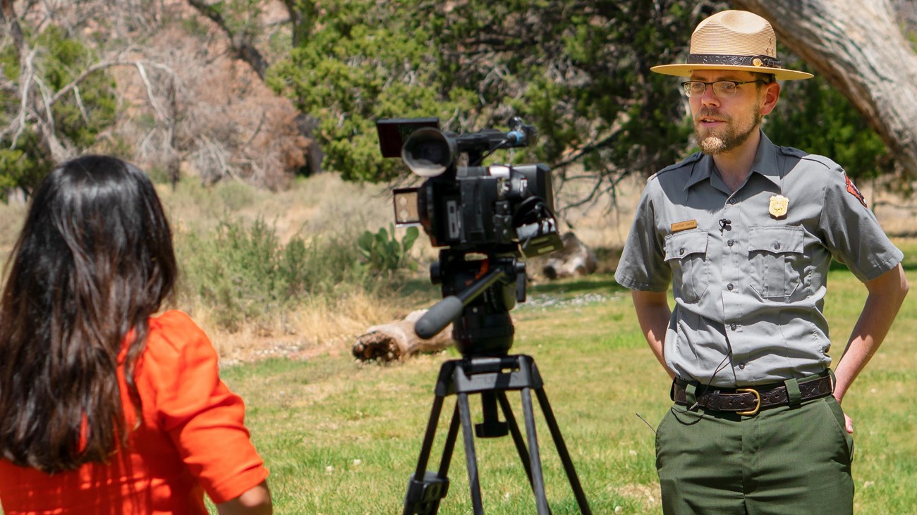 A woman in an orange shirt interviews a park ranger in a grass field.