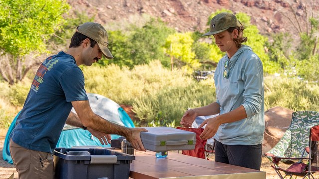 Two people unpack food at a picnic table in a campground.