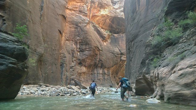 Two hikers walk through a raver between towering walls.