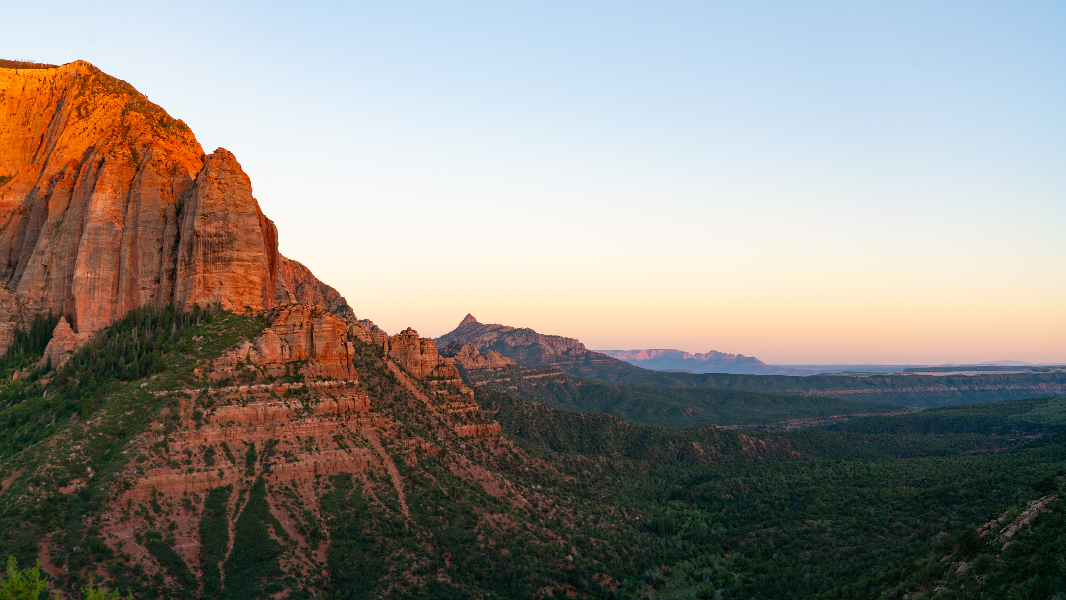 A vast wilderness area at sunset. Tall sandstone peaks stand over valleys and ridges of green trees.