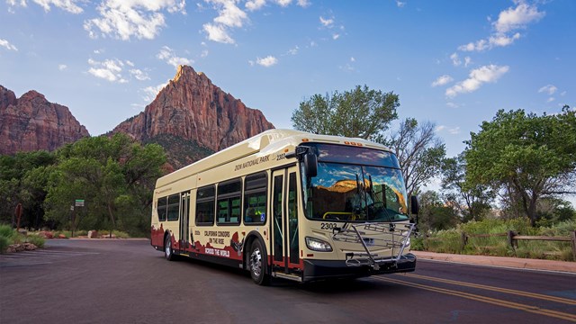 A Zion Canyon shuttle on a red road, with red sandstone formations in the background.