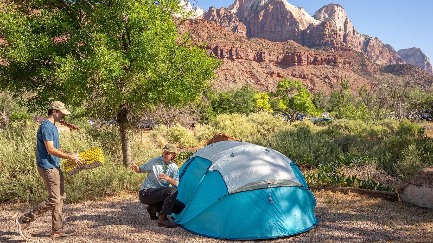 A blue tent on the ground next with two people setting up camp. Sandstone cliffs in background.