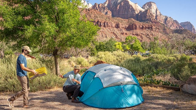 A blue tent on the ground next with two people setting up camp. Sandstone cliffs in background.